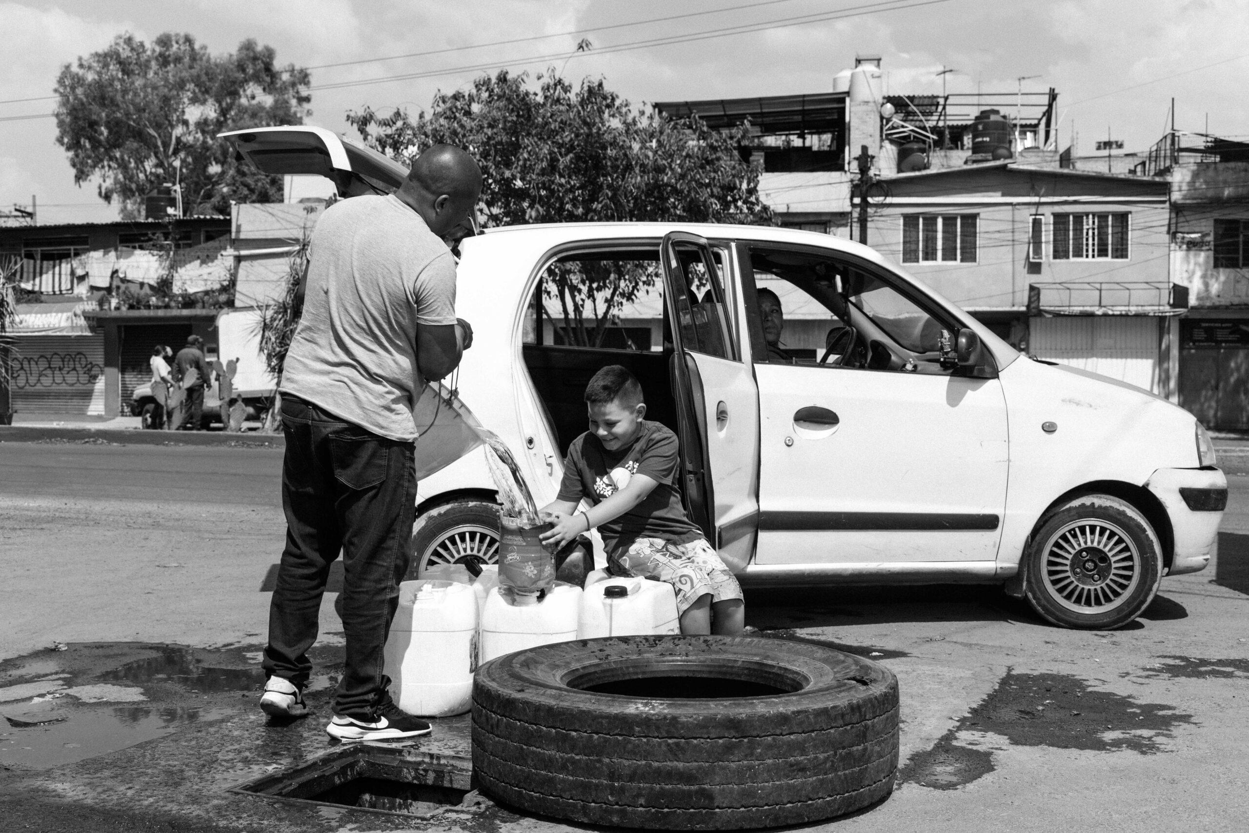 A young boy helps his father fill a can of water that he has just drawn from a manhole in the street, May 27, 2024.