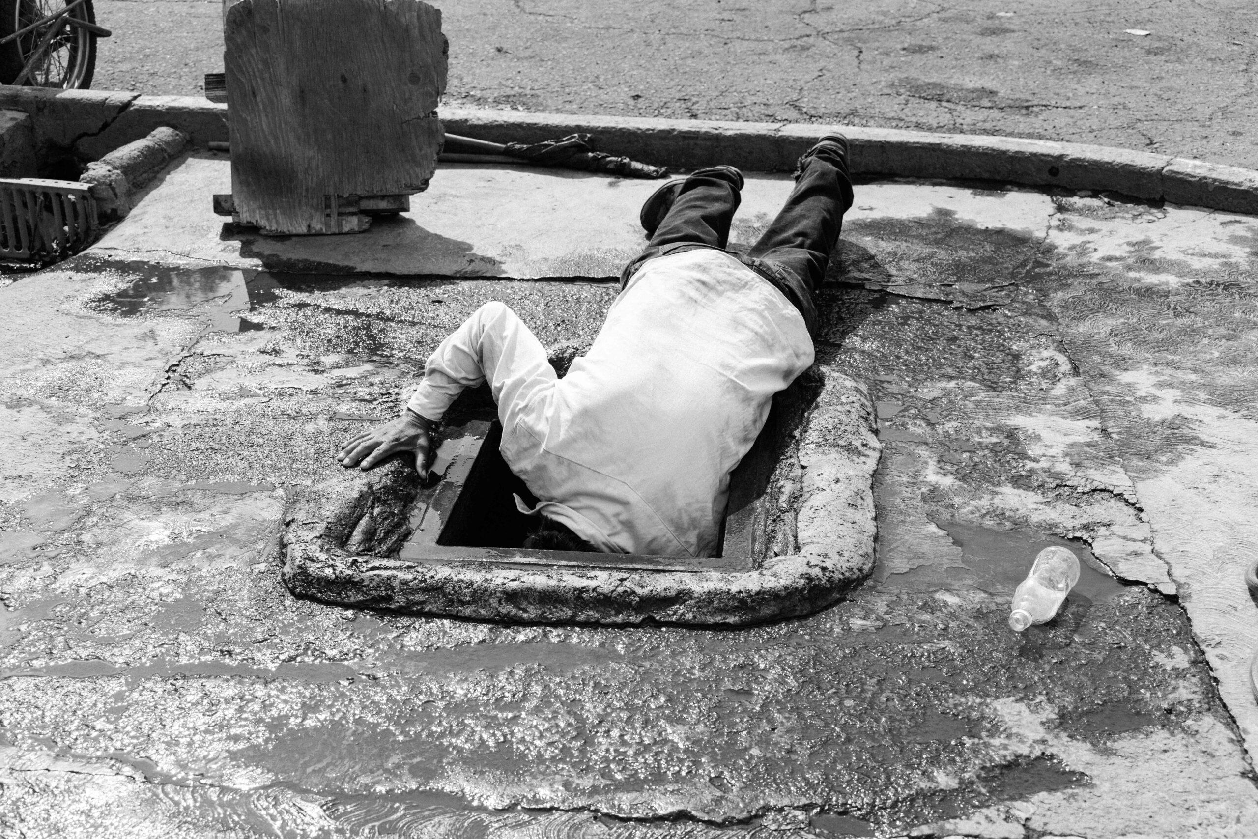 A man sticks his arm and head into a manhole to extract water with a bucket, May 27, 2024.
